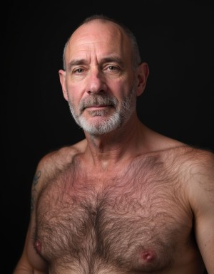Mature shirtless man portrayed from the front against a black background, with soft studio lighting that highlights his face, shoulders, and bare torso. He has a partially bald head, short gray hair on the sides, a trimmed gray-and-white beard, pronounced eyebrows, and a serious, calm expression while looking directly at the camera. His chest is covered with abundant hair, with visible nipples and natural skin without obvious retouching; a dark tattoo is partially visible on his left arm. The intimate portrait-style composition emphasizes the texture of the skin, relaxed musculature, age, masculine presence, and dramatic contrast with the dark background.