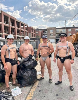 Four men pose smiling on a street next to a construction area, dressed in very revealing fetish-uniform-style outfits: tight suspenders over bare torsos, minimal jockstrap- or brief-like garments covering the crotch, black belts, and work boots. Two wear white construction helmets, another a blue cap, and the one on the right a black police cap, reinforcing a worker-and-officer aesthetic. Around them are large black garbage bags, debris on the ground, containers, tires, and machinery, with brick buildings under construction in the background. The scene has bright daylight, a partly cloudy sky, and an urban industrial atmosphere, blending a humorous pose, muscular physiques, and provocative clothing.