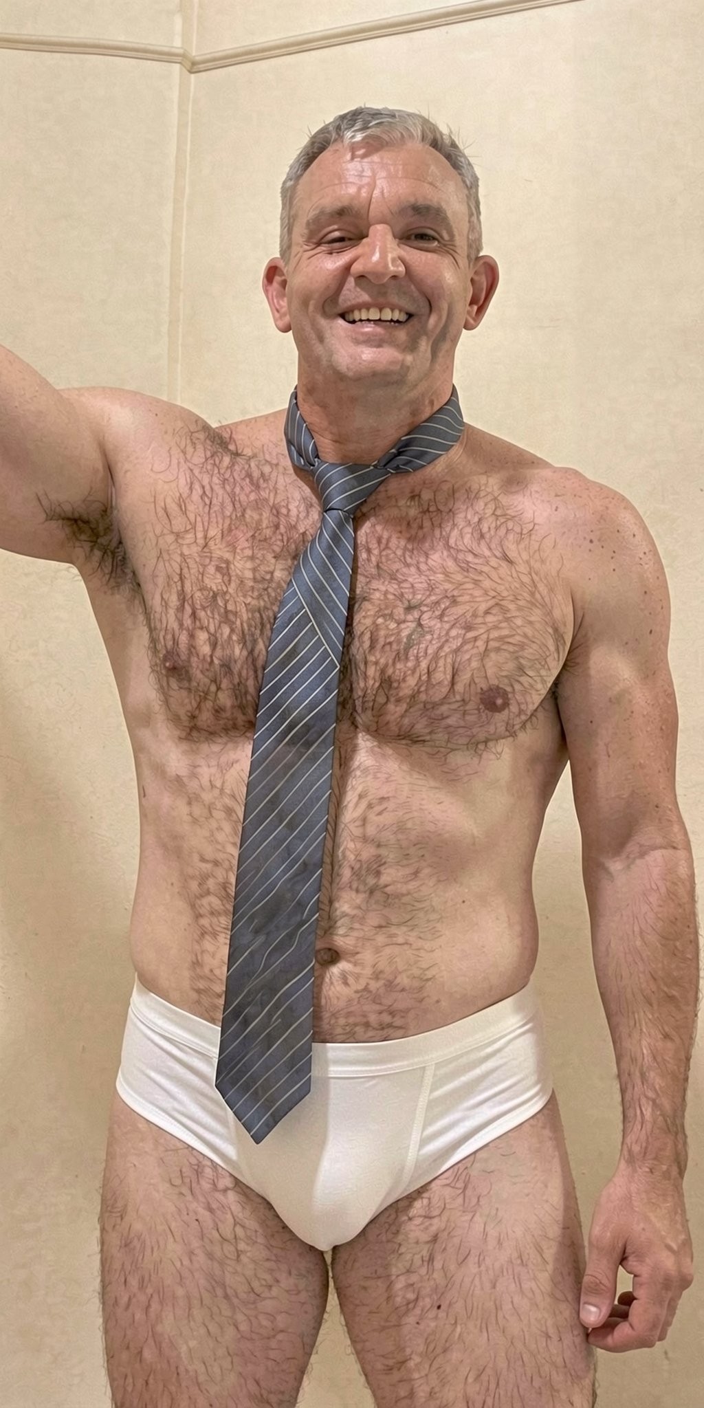 Mature man with an athletic, muscular body poses smiling in a bathroom with light-colored tiles, wearing only tight white briefs and a gray striped tie over his bare torso. He has short gray hair, a mustache, abundant hair on his chest, abdomen, armpits, and legs, and raises one arm as if taking a photo in front of the mirror. The underwear shows a discreet front bulge, while the relaxed and confident posture highlights the broad shoulders, defined arms, and masculine look of the scene, with warm lighting and a simple beige wall background.