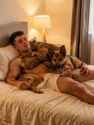 Un hombre sin camiseta descansa tumbado en una cama deshecha con sábanas beige, apoyado sobre almohadas y con unos pantalones cortos claros, mientras abraza a un perro grande de pelaje marrón y negro que está recostado sobre su torso. El perro, similar a un pastor, mira hacia la cámara con la lengua fuera y expresión tranquila, creando una escena íntima y relajada de compañía en un dormitorio. La luz cálida de una lámpara de mesa ilumina suavemente la habitación, junto a un cabecero tapizado, cortinas grises y una ventana lateral. Los tatuajes del hombre, su postura relajada y el ambiente acogedor refuerzan una sensación doméstica, serena y afectuosa.