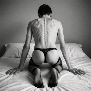 Thin person kneeling on a white bed, seen from behind in a black-and-white photograph of an intimate and artistic tone. The bare back appears very marked, with the spine, ribs, shoulder blades and shoulders defined under a soft light that highlights the texture of the skin. They wear a black lace thong that leaves the buttocks exposed and dark fishnet stockings up to the thighs, while resting both open hands on the wrinkled sheet. The scene takes place in a simple bedroom, with white pillows and a plain gray wall in the background, creating a minimalist, sensual and vulnerable composition.