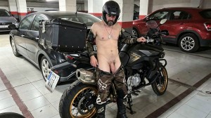 Un hombre con casco negro y botas de motorista posa en un garaje junto a una motocicleta negra de estilo touring o adventure, con maletas laterales y baúl trasero, mientras lleva los pantalones de cuero bajados hasta los muslos, dejando al descubierto el torso desnudo, el abdomen, el pene y los testículos. Apoya una mano en el manillar de la moto y mantiene una actitud desafiante o provocadora, con colgante al cuello y guantes oscuros. Alrededor hay coches aparcados, incluido un vehículo rojo al fondo y una berlina negra a la izquierda, en un aparcamiento cubierto con suelo de baldosas, columnas blancas y franjas rojas.