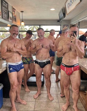 Cinco hombres sonrientes posan en un local tipo bar o cafetería junto a una encimera de piedra, descalzos y vestidos únicamente con slips o bañadores ajustados de estilo deportivo, varios con el logotipo de Adidas visible. El ambiente es informal y desenfadado, con cuerpos velludos, torsos desnudos, piernas descubiertas y paquetes marcados bajo la ropa interior; uno de ellos sostiene un móvil frente a un espejo para hacer la foto, mientras otros ríen y charlan relajadamente. Al fondo se ven más clientes, una barra, carteles de menú, ventanas con palmeras en el exterior y una decoración sencilla de azulejos y madera, creando una escena social, veraniega y de vestuario improvisado.