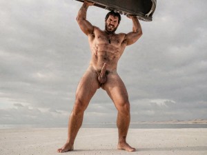 Un hombre desnudo y muy musculoso posa de pie sobre una llanura clara y desértica, con las piernas abiertas y los brazos levantados sosteniendo por encima de la cabeza una gran pieza oscura que parece una tabla o estructura metálica. Su cuerpo aparece completamente expuesto, con pecho, abdomen marcado, muslos fuertes y pene visible, transmitiendo una estética de fuerza física y resistencia. Tiene barba, pelo oscuro despeinado y una expresión intensa, casi desafiante, mientras mira hacia delante. El fondo muestra un paisaje amplio y vacío, parecido a un salar o playa seca, bajo un cielo gris cubierto de nubes, creando una atmósfera dramática, cruda y cinematográfica.