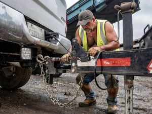 A worker wearing a yellow reflective vest, cap, and gloves handles the trailer hitch next to the rear of a white pickup truck in a muddy outdoor heavy-duty work area. The scene is notable for the metal coupling, the hanging safety chains, the trailer wiring, and the black structure of the tongue, with red reflective markings and signs of wear from use. The man appears focused, leaning over the mechanism, wearing work clothes, dirty boots, and looking like a construction or transport worker, while adjusting the connection between the vehicle and the trailer in an industrial or construction-site environment.