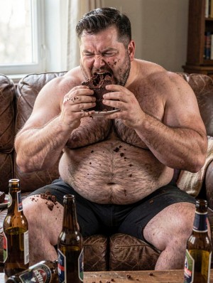 Un hombre corpulento y sin camiseta está sentado en un sofá de cuero marrón, comiendo con ansia un gran trozo de tarta de chocolate que sostiene con ambas manos sobre un plato. Tiene la cara, la barba y los dedos manchados de chocolate, con migas esparcidas por el pecho velludo, el abdomen, los muslos y el asiento, reforzando una escena desordenada y compulsiva. Lleva pantalones cortos oscuros y aparece rodeado de varias botellas de cerveza abiertas colocadas en primer plano sobre una mesa baja, mientras la luz natural entra por una ventana lateral y una estantería al fondo sugiere un salón doméstico descuidado.
