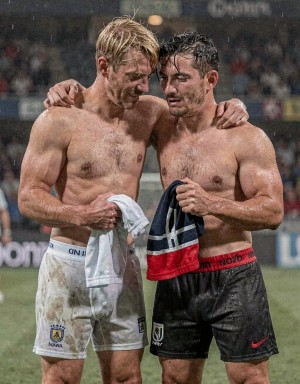 Dos futbolistas empapados y sin camiseta se abrazan sobre el césped tras un partido bajo la lluvia, con el estadio y el público desenfocados al fondo. El jugador de la izquierda, rubio, lleva pantalón corto blanco manchado de barro con el escudo de Suecia y sostiene una camiseta blanca en la mano; el de la derecha, moreno y con barba corta, viste pantalón negro mojado con detalles rojos y agarra una prenda oscura con franjas roja y blanca. Ambos aparecen exhaustos, sudorosos y cubiertos de agua, transmitiendo compañerismo, cansancio y emoción después de un encuentro intenso de fútbol.