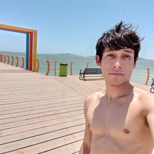 Un hombre joven aparece en primer plano haciéndose un selfi en un paseo marítimo de madera, sin camiseta y bajo una luz intensa de día soleado. Tiene el pelo oscuro despeinado por el viento, expresión tranquila y mirada directa a la cámara. Al fondo se ve el mar en calma de color turquesa, una barandilla naranja que bordea el muelle, bancos negros, una papelera verde y una estructura rectangular naranja tipo marco junto al paseo. El cielo está despejado y el paisaje costero transmite ambiente de verano, playa, vacaciones y aire libre.