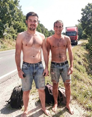Two young men appear standing by the shoulder of a rural road, shirtless and barefoot, wearing worn denim shorts stained with mud, under intense summer light. Both smile while looking at the camera, with bare torsos, short hair, and a tired or adventurous appearance, as if they had come from a hike, an improvised trip, or some outdoor activity. At their feet are black backpacks resting on the gravel and grass, while in the background a red truck can be seen driving along the road lined with leafy trees and dense vegetation, creating a scene of travel, road, nature, and informal rest.
