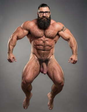 A highly detailed full-body nude photograph of a 40-year-old dominant muscle bear named Hipsterbarba, standing confidently in a well-lit studio with dramatic lighting that accentuates every muscle. He is 180cm tall, 80kg with 8% body fat, extremely muscular and symmetrical. Massive rounded hairy pecs with clear separation and dark pointed nipples, shredded six-pack abs with deep cuts and visible obliques forming a strong V-taper, wide shoulders, vascular arms, huge quads and defined legs. Very dense black masculine body hair covering his chest, running down in a thick treasure trail to a very bushy dark pubic area. He has a perfectly groomed long Bandholz beard that reaches mid-chest, thick mustache with curled upward spiral ends, modern high fade undercut hairstyle, black hipster glasses, and a small tattoo on his right bicep that reads 