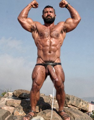 A hyper-masculine ancient Zapotec warrior named Cocijo, 26 years old, standing perfectly still under the brutal midday sun on the rocky summit of Cerro La Cantera in San Pablo Huitzo, Oaxaca, 600 AD. Completely naked except for worn leather strip sandals. Square jaw clenched, thick veiny neck, bronze skin drenched in sweat. Extremely powerful and fully flexed physique: massive square pectorals tensed to the maximum, thick dark wet chest hair matted against the hard muscle, large erect nipples. Deeply carved eight-pack abs glistening with sweat, thick black treasure trail running down to a dense dark pubic bush. Enormous arms raised and flexed hard overhead, biceps bulging like cannonballs with thick veins, triceps perfectly defined and tensed like horseshoes. Thick hairy thighs locked in full tension. His massive 35cm cock is rock-hard and throbbing violently, held tightly at the very base by Gayon Von Neumann’s strong hand, the swollen head continuously leaking thick white precum that runs down the shaft and over the fingers. A thick black leather cock ring is locked brutally tight at the root, behind his heavy balls, making the entire package look painfully swollen, darker and pulsing non-stop. Cocijo stares straight up with total obedience, every muscle in his body still tensed and displayed for inspection. Raw, dominant, extremely virile ancient masculine atmosphere, dramatic sunlight casting strong shadows on every ridge of muscle, hyper-realistic and extremely detailed.