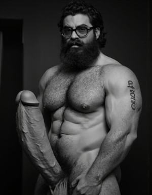 Fotografía hiperrealista en blanco y negro de un hombre adulto musculoso a la vista desde las rodillas para arriba hasta la cabeza, cuerpo muy delgado con musculación marcada y definida, rostro masculino, totalmente desnudo con polla erecta de treinta centímetros de longitud y ocho centímetros de diámetro, tiene rasgos marcados duros expresivos, barba densa poblada tupida oscura estilo hipster cubriendo completamente mandíbula mentón mejillas con grosor uniforme textura abundante muy definida, bigote denso tupido oscuro conectado perfectamente con la barba completa cubriendo completamente el labio superior, pelo oscuro medio peinado completamente recto hacia arriba con volumen considerable estilo hipster moderno quiff puntas verticales texturizadas hacia el cielo con altura prominente, cejas gruesas oscuras pobladas muy marcadas definidas, gafas de montura completa negra cuadradas grandes estilo hipster moderno sobre el puente de la nariz enmarcando unos ojos oscuros intensos que miran directamente a cámara con expresión seria penetrante confiada, expresión facial seria masculina sin sonrisa con mirada directa desafiante, pómulos marcados definidos angulares, nariz recta proporcionada, labios medios apenas visibles completamente cubiertos por bigote denso que conecta con la barba hipster completa, torso desnudo muy musculoso con pectorales enormes desarrollados y definidos, hombros anchos musculosos deltoide prominentes, brazos musculosos gruesos con bíceps y tríceps marcados visibles a ambos lados del cuerpo, abdomen tonificado marcado visible desde la cintura, piel con textura realista detallada poros imperfecciones sutiles, vello corporal denso oscuro abundante cubriendo pectorales abdomen brazos hombros con textura natural masculina muy visible, tatuaje en tinta negra en el bíceps del brazo derecho con la palabra exacta 
