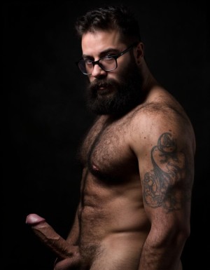 Fotografía hiperrealista de un hombre adulto musculoso a la vista desde las rodillas para arriba hasta la cabeza, rostro masculino idéntico a la imagen de referencia totalmente desnudo con polla erecta de treinta centímetros de longitud y ocho centímetros de diámetro, tiene rasgos marcados duros expresivos, barba densa poblada tupida oscura estilo hipster cubriendo completamente mandíbula mentón mejillas con grosor uniforme textura abundante muy definida, bigote denso tupido oscuro conectado perfectamente con la barba completa cubriendo completamente el labio superior, pelo oscuro medio peinado completamente recto hacia arriba con volumen considerable estilo hipster moderno quiff puntas verticales texturizadas hacia el cielo con altura prominente, cejas gruesas oscuras pobladas muy marcadas definidas, gafas de montura completa negra cuadradas grandes estilo hipster moderno sobre el puente de la nariz enmarcando unos ojos oscuros intensos que miran directamente a cámara con expresión seria penetrante confiada, expresión facial seria masculina sin sonrisa con mirada directa desafiante, pómulos marcados definidos angulares, nariz recta proporcionada, labios medios apenas visibles completamente cubiertos por bigote denso que conecta con la barba hipster completa, torso desnudo muy musculoso con pectorales enormes desarrollados y definidos y velludos peludos, hombros anchos musculosos deltoide prominentes, brazos musculosos con bíceps y tríceps marcados visibles a ambos lados del cuerpo, abdomen tonificado marcado visible desde la cintura, piel con textura realista detallada poros imperfecciones sutiles, vello corporal denso oscuro abundante cubriendo pectorales abdomen brazos hombros con textura natural masculina muy visible, tatuaje en tinta negra en el bíceps del brazo derecho con la palabra exacta a la vista: 