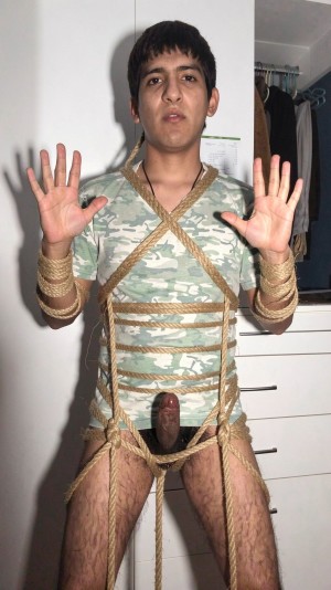 Un hombre joven aparece de pie frente a una pared blanca y un armario abierto con ropa colgada, mirando directamente a la cámara con las manos levantadas y las palmas abiertas. Lleva una camiseta verde de camuflaje y está parcialmente desnudo de cintura para abajo, con el pene expuesto entre las piernas. Varias cuerdas gruesas de color beige rodean su torso, cruzan el pecho, la cintura, las muñecas y los muslos en un atado tipo bondage, con nudos y vueltas visibles que sujetan también la zona de las caderas. La escena tiene iluminación directa de flash, sombras marcadas y un encuadre vertical de habitación doméstica.