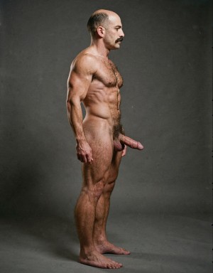 Mike Morris, a 32-year-old extremely lean Caucasian male with only 8% body fat, stands completely naked in a rigid military attention pose captured in a full-body exact side profile at 90 degrees from his right side. The shot runs uninterrupted from the tips of his size 44 slightly hairy feet all the way up to the top of his bald head, showing his entire body without any cropping whatsoever. His physique is dry, highly vascular and powerfully defined: massive square pectorals seen in perfect side silhouette, dense dark brown chest hair forming a thick T-pattern that continues across his shoulders and back, sharply carved six-pack abs in clear profile, narrow waist, wide flaring lats, powerful thighs and diamond-shaped calves covered in the same dark hair. Dramatic directional studio lighting falls from high and slightly in front of him, creating strong rim lighting that traces every sharp edge of his muscular outline, accentuating the texture of his dense dark body hair on chest, shoulders, lower back, ass and legs, while producing deep shadows that highlight every muscle separation, vein and the extreme dryness of his 8% body fat. His advanced Norwood III-IV bald scalp is clearly visible under the light, with very short dense dark brown hair only on the sides and back of his head. His square, angular face is shown in pure right profile: strong jawline, prominent chin with slight central dimple, thick well-groomed dark handlebar mustache, heavy brow and intense deep-set brown eye staring straight forward with a serious, commanding expression. His penis is fully and clearly visible in perfect side view, completely natural and anatomically correct: thick, 24 cm long when fully erect, with a natural slight upward curve, prominent dorsal veins running along the shaft, long foreskin that partially covers the pink-red glans, and noticeably lighter skin tone than the rest of his bronzed body. Below it hang his large, low-hanging testicles with wrinkled darker skin and an extremely dense, curly dark brown pubic bush that climbs almost to his navel and surrounds the base of his cock and balls generously. Every detail from his veined hands and forearms to his muscular ass, hamstrings, diamond calves and hairy toes is perfectly proportioned and free of any deformation. Photorealistic, high-contrast, masculine 1970s Colt Studio aesthetic with subtle grainy film texture.
