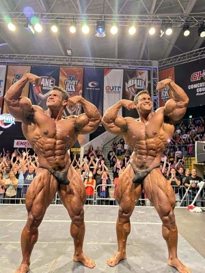 Dos culturistas muy musculados posan sobre un escenario de competición haciendo la pose de doble bíceps frontal, con el torso desnudo, bañador de posing oscuro y la piel bronceada y aceitada para resaltar la definición extrema de abdominales, pectorales, brazos, hombros y piernas. Ambos sonríen bajo una intensa iluminación de focos, mostrando una vascularidad marcada y una simetría corporal propia del culturismo profesional. Al fondo aparece un público numeroso detrás de vallas, con personas aplaudiendo y grabando, además de pancartas y logotipos de patrocinadores en una feria o campeonato fitness cubierto.