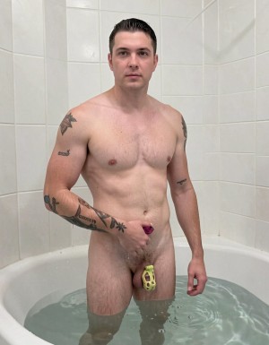 Hombre desnudo de pie dentro de una bañera blanca con agua hasta los muslos, en un baño de azulejos claros, mirando directamente a la cámara con expresión neutra y el pelo corto peinado hacia atrás. Tiene complexión atlética, pecho descubierto con vello corporal y varios tatuajes negros en hombros, brazos y antebrazo. Su pene está cubierto por una jaula de castidad amarilla, mientras sostiene en la mano una pequeña pieza morada cerca del pubis. La escena tiene iluminación fría y uniforme, con reflejos en el agua y un encuadre frontal que centra el cuerpo, los tatuajes, la bañera y el ambiente íntimo del cuarto de baño.