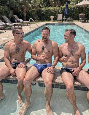 Tres hombres musculosos y sonrientes están sentados en el borde de una piscina al aire libre, con las piernas dentro del agua y bañadores tipo slip en tonos oscuros y azules. Llevan pequeños micrófonos sujetos al pecho o al bañador, lo que sugiere una grabación, entrevista o sesión de contenido junto a la piscina. El ambiente es soleado y relajado, con tumbonas, sombrillas, vegetación abundante y palmeras alrededor, creando un entorno de resort o patio privado. Los hombres, de pelo corto y barba o vello corporal visible, interactúan entre risas y miradas cómplices, transmitiendo una escena veraniega, informal y social. En el extremo derecho aparece parcialmente recortada otra persona sentada junto a ellos.