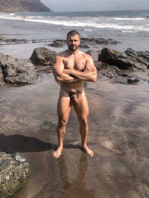 Un hombre desnudo posa de pie en una playa rocosa, con los brazos cruzados sobre el pecho y mirando directamente a la cámara, mostrando el torso velludo, las piernas musculadas y el pene visible. Está descalzo sobre arena húmeda cubierta por una fina lámina de agua, rodeado de grandes rocas oscuras y charcos de marea que reflejan la luz. Al fondo se extiende el mar con olas rompiendo cerca de la orilla, varias personas bañándose o caminando en la distancia y un acantilado costero bajo un cielo despejado, creando una escena natural de costa, surf y paisaje marítimo.