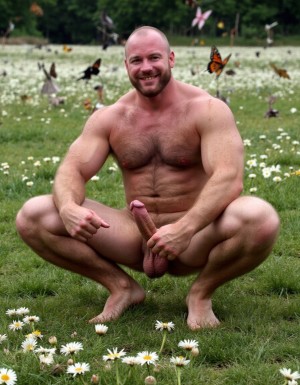 hombre culturista de 35 años posando desnudo de cuerpo completo, sonriente, tensa los bíceps, tiene un pene de 40 cm de longitud y 7 cm de diámetro, tiene grandes dorsales, piernas, tiene cintura estrecha, es un poco calvo, tiene grandes pezones rosas. Tiene un pene de 40 cm de longitud y 7 cm de diámetro. Tiene mucho vello en pecho y brazos y piernas. Tiene testículos de 10 cm de diámetro que están colgando y se mueven al compás de los movimientos del cuerpo. Tiene pies con sus uñas pintadas de color rojo. Fondo de muchas margaritas de todos los colores y miles de mariposas volando. Está agachado en la hierva rodeado de margaritas. Está agachado con las piernas tensadas con los pies hacia arriba para enseñar su ano. Tiene el ano relajado y abierto y a la vista del espectador. Por su pene erecto.