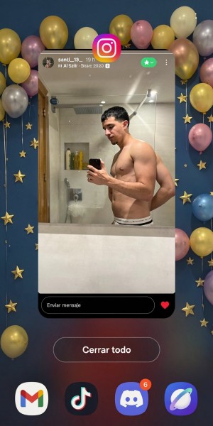 Screenshot of a mobile phone with an Instagram story open, showing a shirtless man taking a selfie in front of a modern bathroom mirror. He is in profile, holding a black smartphone with one hand and placing the other behind his back, with his bare torso visible and wearing light-colored pants with an elastic waistband. The bathroom has a white sink in the foreground, a large mirror, ceiling lighting, a shower with a glass screen, and a light-colored wall with a clean, minimalist look. Around the story there is a decorative background of gold, pink, and blue balloons with hanging stars on a dark blue background. At the bottom appear the “Send message” bar, a red heart, the “Close all” button, and several app icons such as Gmail, TikTok, and Discord.