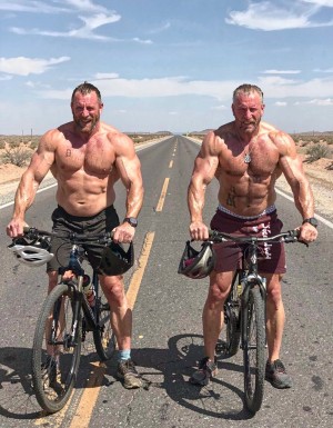 tres hombres muy viejos de 80 años  musculosos semidesnudos, de aspecto agresivo, malotes macarras, con los  penes visibles, torsos desnudos, se han quitado el traje  de ciclista, muestran pechos y abdómenes hipermusculosos, junto a sus bicis, cascos cascos en sus manos, el fondo una carretera secundaria perdida en desierto de los Monegros. 