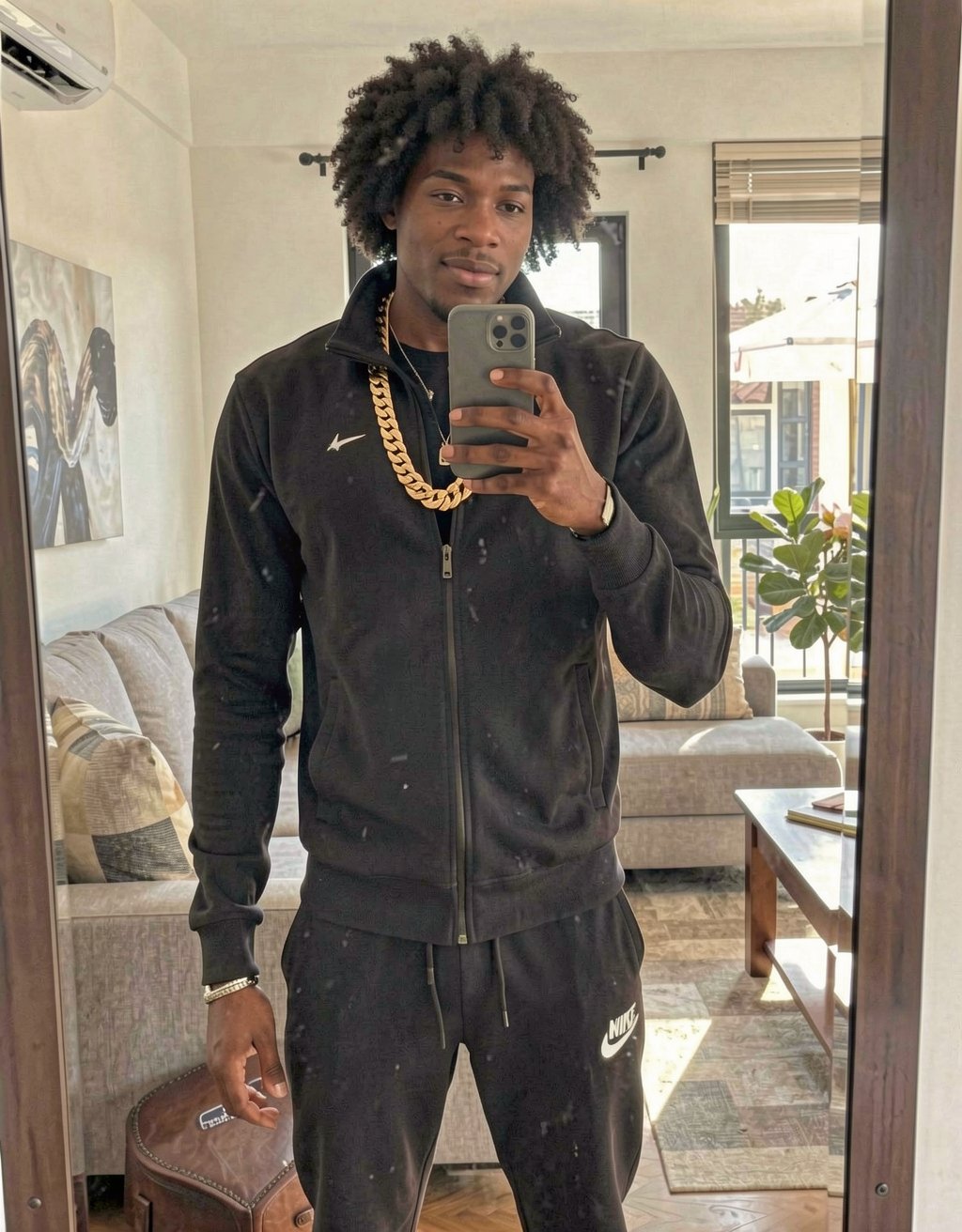 A tall, muscular 20-year-old Moroccan guy standing in a modern living room, taking a mirror selfie. He has a shaved head, sharp Arab features, dark intense eyes, thick eyebrows, wearing a black Nike tracksuit with gold chains around his neck, looking cocky and arrogant with a slight smirk. Strong jawline, athletic build visible under the clothes, dominant street vibe, realistic photo style, natural indoor lighting.