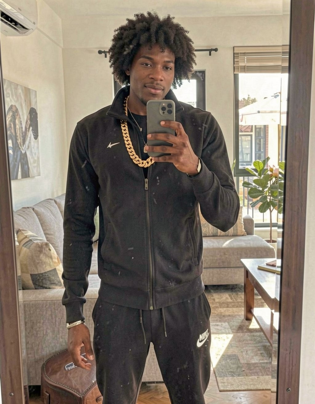 A tall, muscular 20-year-old Moroccan guy standing in a modern living room, taking a mirror selfie. He has a shaved head, sharp Arab features, dark intense eyes, thick eyebrows, wearing a black Nike tracksuit with gold chains around his neck, looking cocky and arrogant with a slight smirk. Strong jawline, athletic build visible under the clothes, dominant street vibe, realistic photo style, natural indoor lighting.