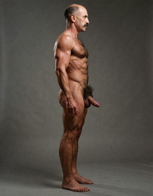 Mike Morris, a 32-year-old extremely lean Caucasian male with only 8% body fat, stands completely naked in a rigid military attention pose captured in a full-body exact side profile at 90 degrees from his right side. The shot runs uninterrupted from the tips of his size 44 slightly hairy feet all the way up to the top of his bald head, showing his entire body without any cropping whatsoever. His physique is dry, highly vascular and powerfully defined: massive square pectorals seen in perfect side silhouette, dense dark brown chest hair forming a thick T-pattern that continues across his shoulders and back, sharply carved six-pack abs in clear profile, narrow waist, wide flaring lats, powerful thighs and diamond-shaped calves covered in the same dark hair. Dramatic directional studio lighting falls from high and slightly in front of him, creating strong rim lighting that traces every sharp edge of his muscular outline, accentuating the texture of his dense dark body hair on chest, shoulders, lower back, ass and legs, while producing deep shadows that highlight every muscle separation, vein and the extreme dryness of his 8% body fat. His advanced Norwood III-IV bald scalp is clearly visible under the light, with very short dense dark brown hair only on the sides and back of his head. His square, angular face is shown in pure right profile: strong jawline, prominent chin with slight central dimple, thick well-groomed dark handlebar mustache, heavy brow and intense deep-set brown eye staring straight forward with a serious, commanding expression. His penis is fully and clearly visible in perfect side view, completely natural and anatomically correct: thick, 24 cm long when fully erect, with a natural slight upward curve, prominent dorsal veins running along the shaft, long foreskin that partially covers the pink-red glans, and noticeably lighter skin tone than the rest of his bronzed body. Below it hang his large, low-hanging testicles with wrinkled darker skin and an extremely dense, curly dark brown pubic bush that climbs almost to his navel and surrounds the base of his cock and balls generously. Every detail from his veined hands and forearms to his muscular ass, hamstrings, diamond calves and hairy toes is perfectly proportioned and free of any deformation. Photorealistic, high-contrast, masculine 1970s Colt Studio aesthetic with subtle grainy film texture.