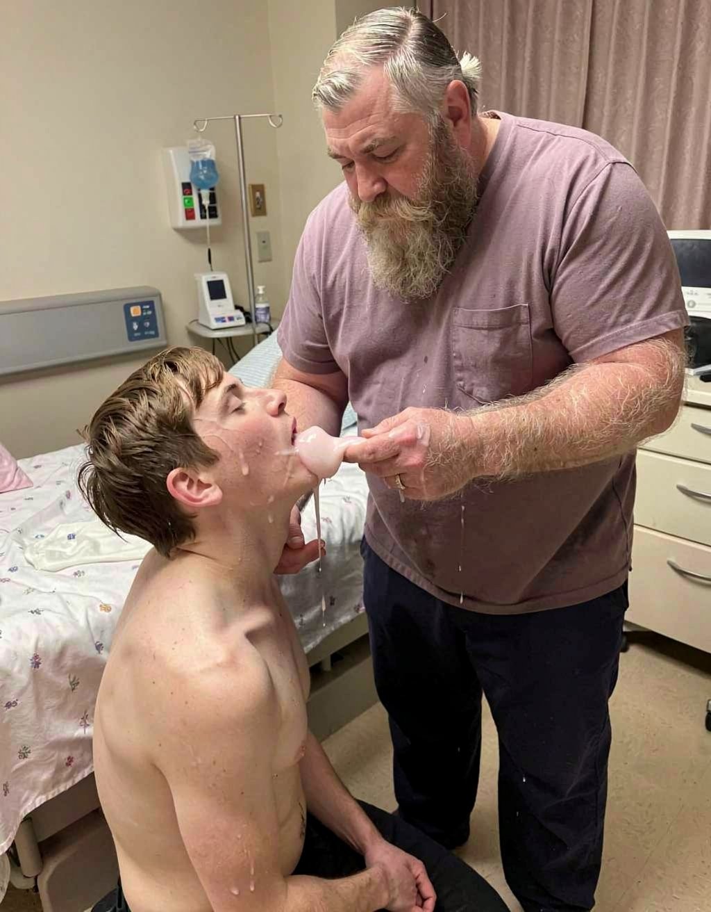 Un hombre mayor con barba gris y camiseta malva atiende de cerca a un joven sin camiseta sentado en una habitación de hospital o consulta clínica, inclinándole suavemente la cabeza mientras introduce un dedo en su boca o revisa la zona de la lengua y la barbilla. El joven permanece sentado junto a una cama médica con sábanas estampadas, con líquido blanquecino escurriéndole desde la boca por el cuello y el pecho, lo que sugiere una revisión, limpieza o procedimiento asistido. Al fondo aparecen equipamiento sanitario, dispensadores, cortinas beige, una mesilla con cajones y paredes claras, reforzando el contexto médico y de cuidado personal.