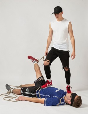 A man is lying on his back against a white studio background, with several ropes wrapped around his torso, arms, and legs, while another man stands beside him in a dominant pose, resting a red sneaker on his chest. The man on the floor is wearing a blue sports shirt, black shorts, gray socks, dark glasses, and appears immobilized, with one leg lifted by a rope that the standing man is holding in his hand. The second man is wearing a black cap, a white sleeveless shirt, tight ripped black pants, and red sneakers, creating a scene of control, submission, and visual tension with an urban and minimalist aesthetic.