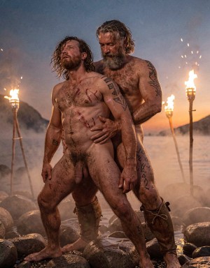 Amleth y Aurvandil.  Versión cinematográfica explícita de la imagen: Dos guerreros vikingos adultos de cuerpo entero se aferran en una pose ritual intensa sobre la orilla rocosa de un fiordo nórdico al anochecer, con el guerrero de 29 años delante arqueando la cabeza hacia atrás y el rey guerrero de 48 años sujetándolo por la cintura desde atrás. La niebla baja se arrastra entre piedras húmedas, antorchas reales lanzan chispas y humo ondulante, y la piel muestra poros, barro agrietado, sangre seca, cicatrices elevadas, tatuajes rúnicos oscuros, vello corporal, cabello enmarañado y barbas sucias. El contraluz crepuscular profundo dibuja un degradado de azul cobalto a naranja quemado, mientras una iluminación cinematográfica de tres puntos añade clave alta controlada y relleno lateral cálido sobre los torsos. La composición vertical 9:16 usa lente anamórfica de 40mm, grano analógico tipo Kodak Vision3 500T, contraste HDR con sombras profundas y altas luces contenidas, en un realismo brutal de saga nórdica.