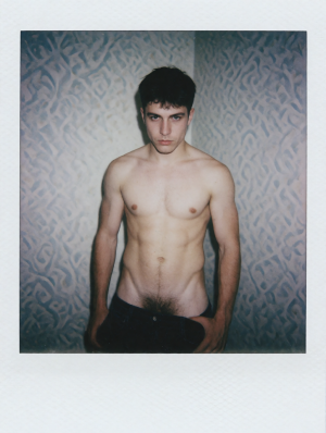 Retrato tipo Polaroid de un hombre joven sin camiseta, de pie frente a una pared con textura azulada y motivos abstractos en relieve, iluminado con un flash directo que resalta el torso desnudo, los hombros, el abdomen y el vello que desciende hacia la cintura. Lleva pantalón oscuro parcialmente bajado o abierto en la zona superior, con una mano apoyada cerca de la cadera, y mantiene una expresión seria e intensa mirando fijamente a la cámara. El encuadre vertical, el borde blanco de la fotografía instantánea y la luz fría crean una estética íntima, cruda y ligeramente melancólica, centrada en el cuerpo masculino, la piel pálida y la pose frontal.