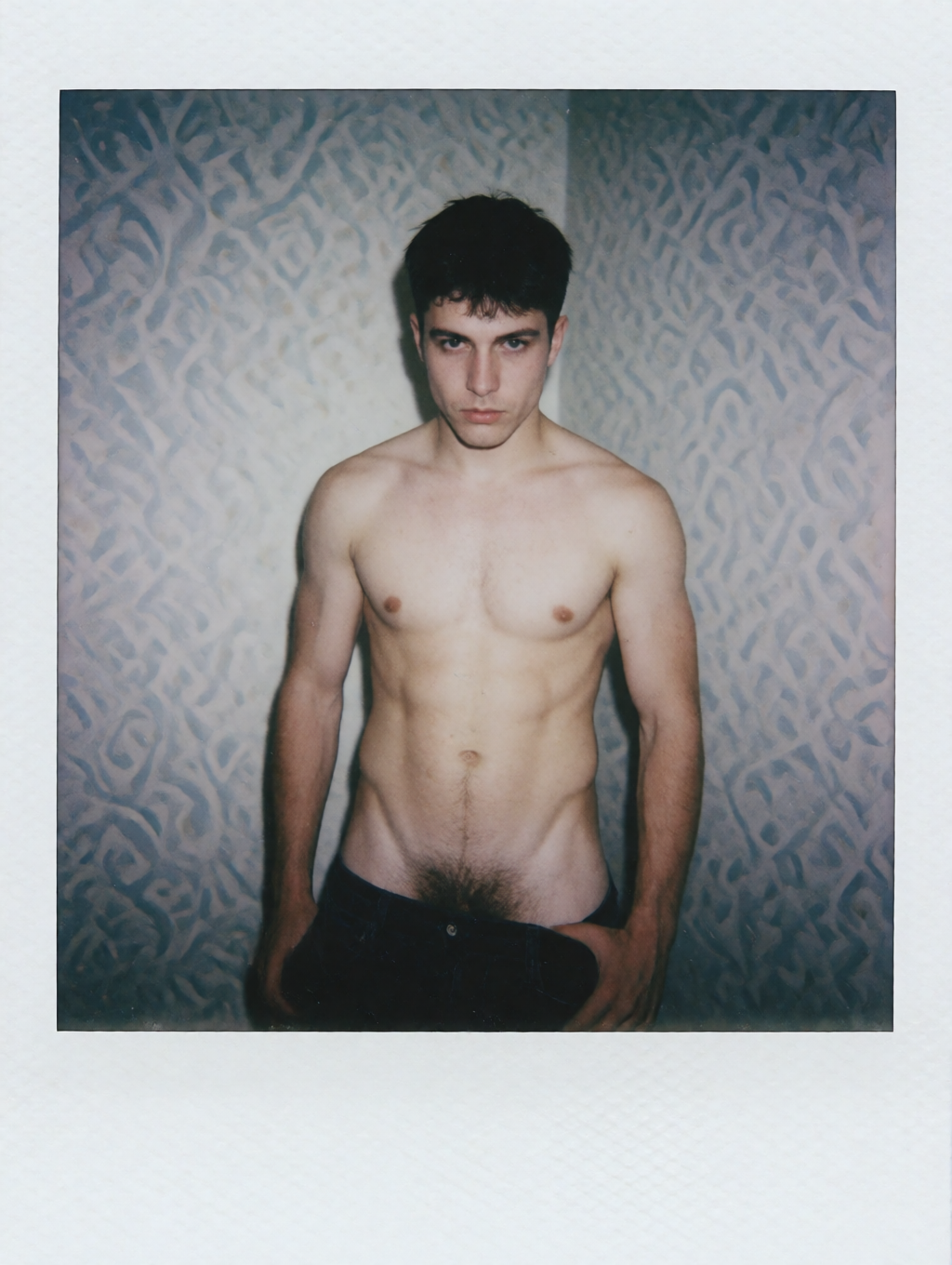 Polaroid-style portrait of a young man without a shirt, standing in front of a wall with a bluish texture and abstract motifs in relief, lit with a direct flash that highlights the bare torso, the shoulders, the abdomen and the hair that descends toward the waist. He wears dark pants partially lowered or open at the upper area, with one hand resting near the hip, and maintains a serious, intense expression staring fixedly at the camera. The vertical framing, the white border of the instant photograph and the cold light create an intimate, raw and slightly melancholic aesthetic, centered on the male body, the pale skin and the frontal pose.
