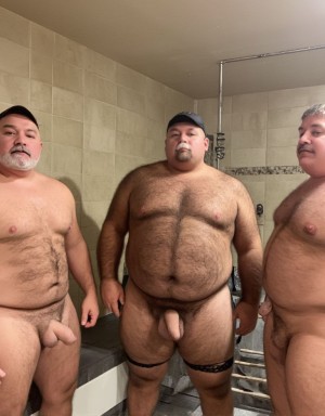 Three naked men pose standing in a beige-tiled bathroom, with their torsos exposed and their penises visible, looking at the camera with neutral expressions. The man in the center, of a very heavy build, wears a black cap and black straps or garters around his thighs; he has abundant hair on his chest and abdomen. On both sides are two other robust men, also with body hair and a beard or mustache, one partially cropped by the right edge. The setting includes a built-in bathtub or bench, a shower with exposed metal piping, and a stepladder leaning in the background, giving the appearance of a spacious bathroom or shower area under renovation.