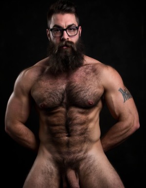 Fotografía hiperrealista de un hombre adulto musculoso a la vista desde las rodillas para arriba hasta la cabeza, rostro masculino idéntico a la imagen de referencia totalmente desnudo con polla erecta de treinta centímetros de longitud y ocho centímetros de diámetro, tiene rasgos marcados duros expresivos, barba densa poblada tupida oscura estilo hipster cubriendo completamente mandíbula mentón mejillas con grosor uniforme textura abundante muy definida, bigote denso tupido oscuro conectado perfectamente con la barba completa cubriendo completamente el labio superior, pelo oscuro medio peinado completamente recto hacia arriba con volumen considerable estilo hipster moderno quiff puntas verticales texturizadas hacia el cielo con altura prominente, cejas gruesas oscuras pobladas muy marcadas definidas, gafas de montura completa negra cuadradas grandes estilo hipster moderno sobre el puente de la nariz enmarcando unos ojos oscuros intensos que miran directamente a cámara con expresión seria penetrante confiada, expresión facial seria masculina sin sonrisa con mirada directa desafiante, pómulos marcados definidos angulares, nariz recta proporcionada, labios medios apenas visibles completamente cubiertos por bigote denso que conecta con la barba hipster completa, torso desnudo muy musculoso con pectorales enormes desarrollados y definidos y velludos peludos, hombros anchos musculosos deltoide prominentes, brazos musculosos gruesos con bíceps y tríceps marcados visibles a ambos lados del cuerpo, abdomen tonificado marcado visible desde la cintura, piel con textura realista detallada poros imperfecciones sutiles, vello corporal denso oscuro abundante cubriendo pectorales abdomen brazos hombros con textura natural masculina muy visible, tatuaje en tinta negra en el bíceps del brazo derecho con la palabra exacta a la vista 
