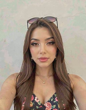 Young woman posing in a close-up selfie, with her face centered and a serene expression in front of a light wall with a worn texture, greenish tones, and subtle cracks. She has long brown hair, loose and styled in soft waves, sunglasses resting on her head, pronounced makeup with defined eyebrows, long eyelashes, eyeliner, highlighter, and pink lips. She wears a black spaghetti-strap top with a floral print in pink and green tones, a V-neckline, a thin necklace with a small pendant, and discreet earrings, creating a polished, feminine, and summery style.