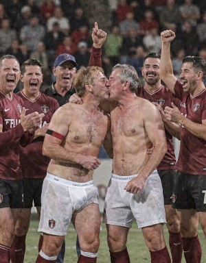 Dos futbolistas celebran efusivamente sobre el césped, sin camiseta y con pantalones blancos bajados hasta quedar en calzoncillos deportivos, besándose en la boca mientras sus compañeros del equipo granate los rodean entre risas, aplausos y gestos de euforia. La escena transmite una celebración intensa de vestuario trasladada al campo, con jugadores levantando los puños, gritando y sonriendo ante la multitud borrosa del estadio al fondo. Los detalles del uniforme, los escudos, las medias granates y la actitud festiva refuerzan el ambiente de fútbol, victoria, camaradería y celebración provocadora.