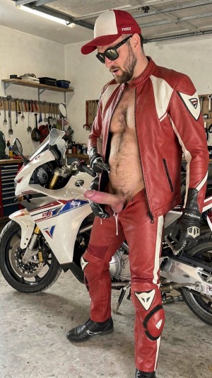 A man with a beard and sunglasses poses in a garage next to a white sport motorcycle, dressed in a partially open red and white leather jumpsuit that leaves his chest and abdomen exposed. He wears a red and white cap, black motorcyclist gloves and boots, with a casual and provocative posture. In the crotch area protrudes a pink penis of artificial appearance, dildo type, creating an explicit and striking scene. In the background appear hanging tools, shelves, a work table and mechanical elements, reinforcing the workshop atmosphere, motorcycling, leather equipment, helmet and sporty biker aesthetic.