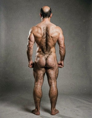 Mike Morris, a 32-year-old extremely lean Caucasian male at only 8% body fat, stands completely naked in a strict military attention pose seen from directly behind in a full posterior view. His back is perfectly straight, shoulders pulled back, arms rigid at his sides with fists clenched, legs straight and together, head held high. The shot captures him from the heels of his feet all the way up to the top of his bald head. His advanced Norwood III-IV baldness is clearly visible from behind, with a smooth tanned scalp on the crown and short dense dark brown hair only on the sides and lower back of the head. His back is highly defined and vascular: wide lats tapering dramatically to a very narrow, tight waist, thick trapezius muscles, and dense dark brown body hair lightly covering the shoulders and becoming much thicker and denser across the lower back and lumbar area, especially between the lats. His ass is tight, muscular and round with almost no fat, the glutes clearly separated. Thick dark hair continues down onto his powerful diamond-shaped calves and the backs of his slightly hairy feet. Dramatic directional studio lighting from above and slightly to the side creates strong highlights on his dry, shredded skin and deep shadows that accentuate every muscle fiber, the texture of his dense back hair, and the extreme vascularity and definition of his 8% body fat physique. Photorealistic, high-contrast, masculine 1970s Colt Studio aesthetic, grainy film texture, commanding and powerful presence.