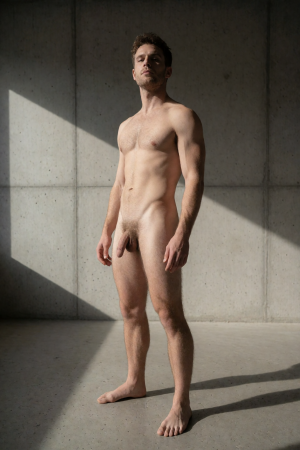 Naked man standing in a minimalist concrete space, with his entire body partially illuminated by a diagonal strip of natural light that casts sharp shadows on the wall and floor. He has a slim athletic build, short brown hair, light beard, and a serious expression, looking toward the camera with an upright and relaxed posture. His torso, abdomen, legs, and penis are visible from the front, without clothing or accessories. The industrial, gray, and austere environment, together with the side lighting, creates a sober studio composition centered on the male figure, anatomy, and the contrast between light and shadow.