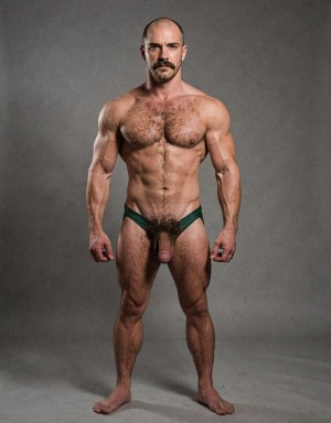 Mike Morris, a 32-year-old Caucasian male with an extremely lean and athletic physique at only 8% body fat, stands completely naked in a strict military attention pose. Full body frontal shot from the tips of his toes to the top of his head. His body is young, dry, and highly defined with sharp muscle separation, visible vascularity, and a very narrow waist. He has advanced Norwood III-IV male pattern baldness with a shiny tanned scalp on top and only short dark brown hair on the sides and back. His face is serious and commanding: square angular jaw, prominent cheekbones, thick dark handlebar mustache, heavy brows, and intense deep-set brown eyes. His massive square pectorals are pushed forward, covered in dense dark brown chest hair that forms a classic thick T-pattern running down the sternum and spreading over his sharply carved six-pack abs. Small dark nipples sit low on the pecs. A prominent trail of dark hair leads to an extremely dense, curly, dark brown pubic bush that climbs almost to his navel. His thick 14 cm flaccid cock hangs heavy over large, low-hanging, hairy testicles, the shaft skin noticeably lighter than his bronzed body. Powerful veined forearms, developed traps, defined quadriceps with clear separation, diamond-shaped calves, and slightly hairy feet complete his figure. He stands perfectly straight, chest out, shoulders back, arms rigid at his sides with fists clenched, head high and gaze fixed forward. Dramatic directional studio lighting from above and slightly to the front creates strong highlights on his dry, vascular skin and deep shadows that accentuate every muscle fiber, the texture of his dense body hair, and the extreme definition of his 8% body fat physique. Photorealistic, high-contrast, 1970s Colt Studio masculine aesthetic, grainy film texture. Tiene un bañador verde oscuro de piscina.