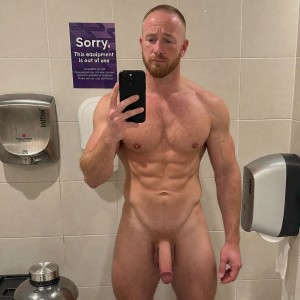Hombre desnudo y musculoso haciéndose un selfi frente al espejo de un baño público, sujetando un móvil negro con una mano mientras mira la pantalla con expresión seria. Tiene el torso definido, abdominales marcados, pecho con vello ligero, barba corta rojiza y el pelo muy rapado. Su pene flácido queda visible entre las piernas, en una postura frontal y estática. El entorno está revestido con azulejos beige y cuenta con dispensadores metálicos y blancos en las paredes, una papelera o contenedor en la parte inferior y un cartel morado que indica que un equipo está fuera de uso, reforzando el contexto de vestuario o aseo público.