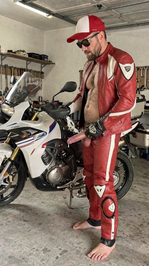 Un hombre con equipación de motorista roja y blanca posa descalzo en un garaje junto a una moto deportiva blanca, con la chaqueta de cuero abierta dejando el pecho y el abdomen al descubierto y los pantalones parcialmente bajados o abiertos, exponiendo el pene de forma explícita. Lleva gorra roja y blanca, gafas de sol oscuras, guantes de moto y pantalones de cuero con protecciones en las rodillas, en un entorno de taller doméstico con herramientas colgadas, estanterías y suelo de hormigón. La escena combina estética motera, garaje mecánico, ropa de cuero, motocicleta deportiva y una pose provocadora de carácter sexual.