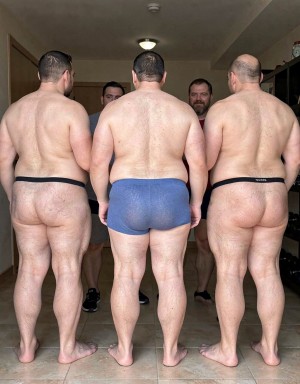 Tres hombres corpulentos aparecen de espaldas en primer plano, descalzos sobre un suelo de baldosas, con la espalda, las piernas y las nalgas descubiertas bajo ropa interior mínima: el de la izquierda lleva un tanga negro estrecho, el del centro unos calzoncillos ajustados azules y el de la derecha otro tanga negro con una pequeña etiqueta visible. Sus cuerpos robustos y velludos ocupan casi toda la escena, iluminada por luz interior en una habitación sencilla con techo bajo, lámpara, puerta lateral y una estantería al fondo. Entre ellos se distinguen parcialmente otros dos hombres de frente, también con poca ropa, creando una composición grupal íntima y directa centrada en cuerpos masculinos, desnudez parcial, traseros expuestos y ambiente doméstico.