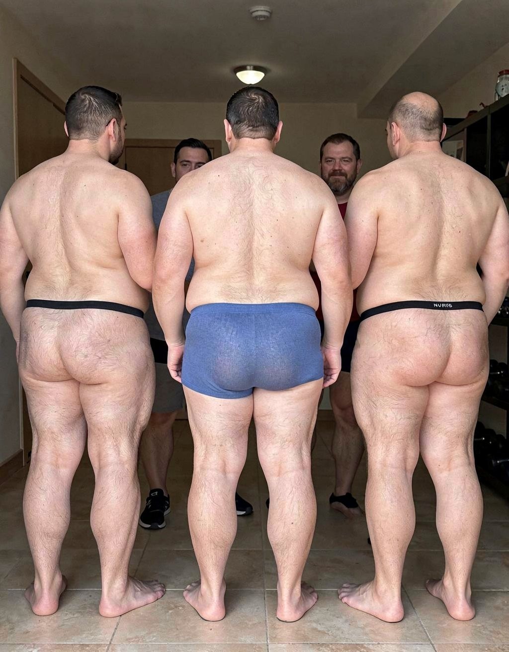 Three burly men appear from behind in the foreground, barefoot on a tiled floor, with their backs, legs, and buttocks uncovered beneath minimal underwear: the one on the left wears a narrow black thong, the one in the center tight blue briefs, and the one on the right another black thong with a small visible label. Their robust, hairy bodies occupy almost the entire scene, lit by indoor light in a simple room with a low ceiling, a lamp, a side door, and a shelf in the background. Between them, two other men can be partially seen from the front, also scantily dressed, creating an intimate and direct group composition centered on male bodies, partial nudity, exposed rears, and a domestic atmosphere.