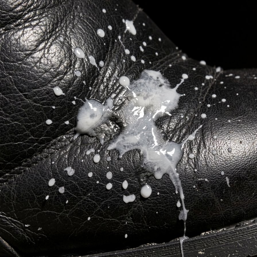 A black leather boot or shoe with a grained texture appears in the foreground, covered with splashes of a thick white liquid that contrasts intensely with the dark, shiny surface. The main stain is concentrated on a fold of the footwear, spreading irregularly with large drops, small scattered spots, and a stream running down toward the lower edge. The lighting highlights the wrinkles of the leather, the satin finish, and the skin-like relief, while the black background keeps all attention on the white splash, the wet dirt, and the detail of the material.