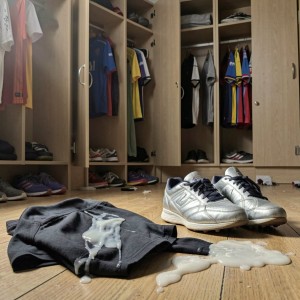 Un vestuario con taquillas de madera abiertas aparece desordenado, con camisetas deportivas colgadas, ropa doblada en los estantes y varias zapatillas repartidas por el suelo. En primer plano hay una sudadera gris tirada sobre el parquet, manchada por un líquido blanco espeso que también se ha derramado formando un charco junto a unas zapatillas plateadas con cordones negros. Al fondo se ven más pares de calzado, prendas de colores y puertas de armario entreabiertas, creando una escena de vestuario deportivo descuidado, con ropa sucia, suelo manchado y ambiente de uso cotidiano tras un entrenamiento.