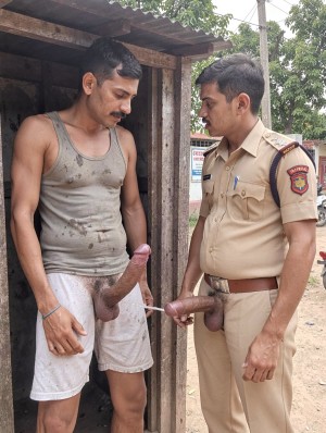 Dos hombres aparecen de pie junto a una pequeña caseta de madera en un entorno exterior, con árboles y suelo de tierra al fondo; uno lleva una camiseta de tirantes gris manchada y pantalón corto blanco, mirando hacia abajo, mientras su pene queda expuesto de forma muy prominente por fuera de la ropa. Frente a él hay un hombre con uniforme caqui de policía, con insignias en las mangas, cinturón marrón y bolígrafo en el bolsillo, que le observa de cerca y también tiene el pene expuesto fuera del pantalón. La escena tiene un aspecto informal y llamativo, con contraste entre la ropa sucia del primer hombre, el uniforme oficial del agente y la desnudez explícita en primer plano.