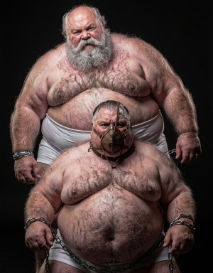 Dos hombres corpulentos y de edad madura posan semidesnudos sobre un fondo negro, iluminados con una luz dura que resalta la piel, el vello corporal abundante, los pliegues del torso y la expresión severa de sus rostros. El hombre del fondo, con barba blanca espesa, calvicie y gesto intimidante, está de pie con el pecho descubierto y ropa interior blanca, llevando grilletes metálicos en las muñecas. Delante aparece otro hombre sentado o agachado, también con el torso desnudo y ropa interior blanca, con una máscara de cuero tipo bozal cubriéndole la boca y correas alrededor de la cabeza; sus muñecas están esposadas y una cadena rodea su cintura. La escena tiene una estética oscura, teatral y de dominación, con elementos de bondage, restricción física y retrato provocador.